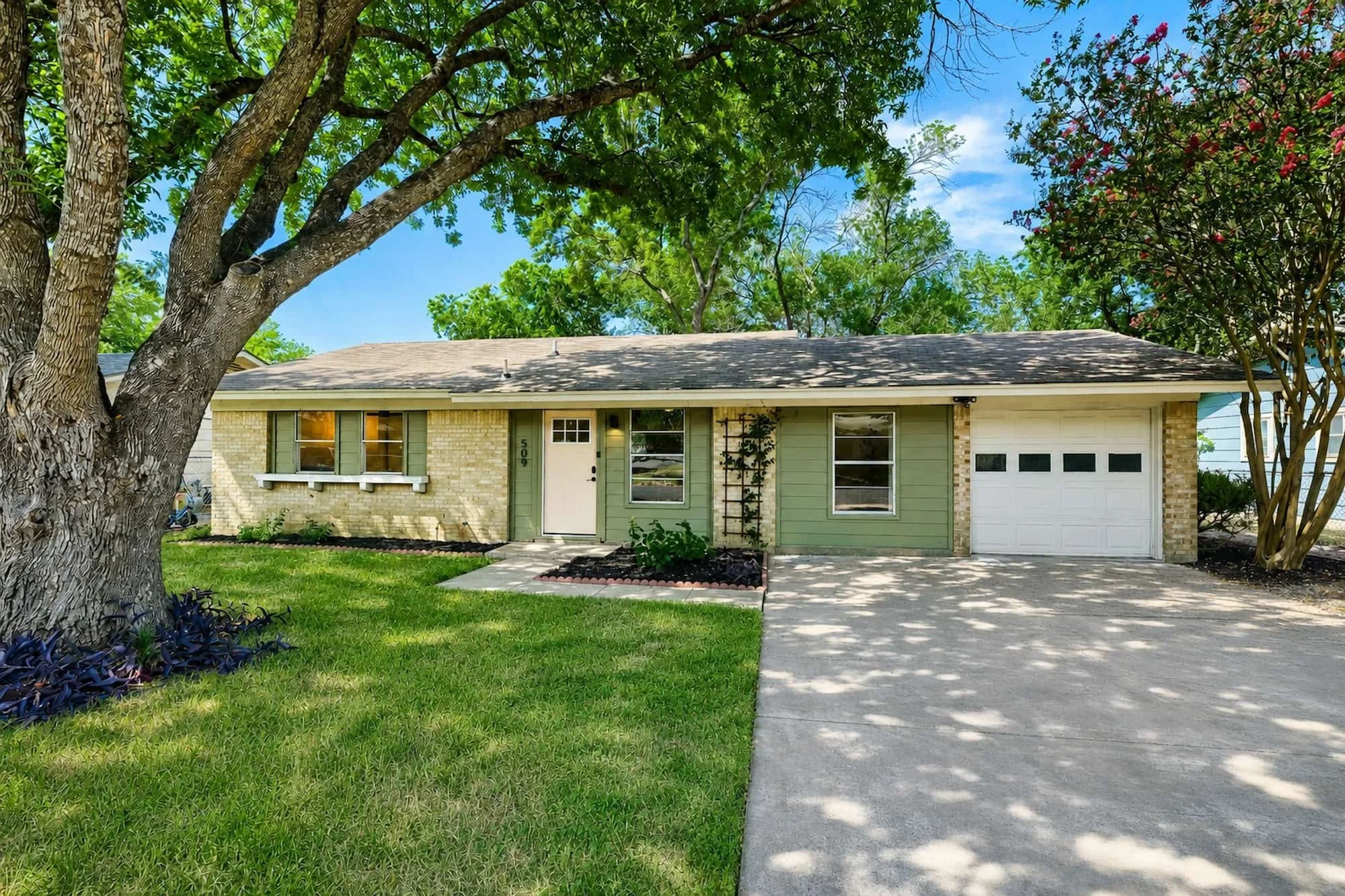 Single-story home with cream brick and sage green siding, an attached garage, and mature shade trees over a green front lawn.