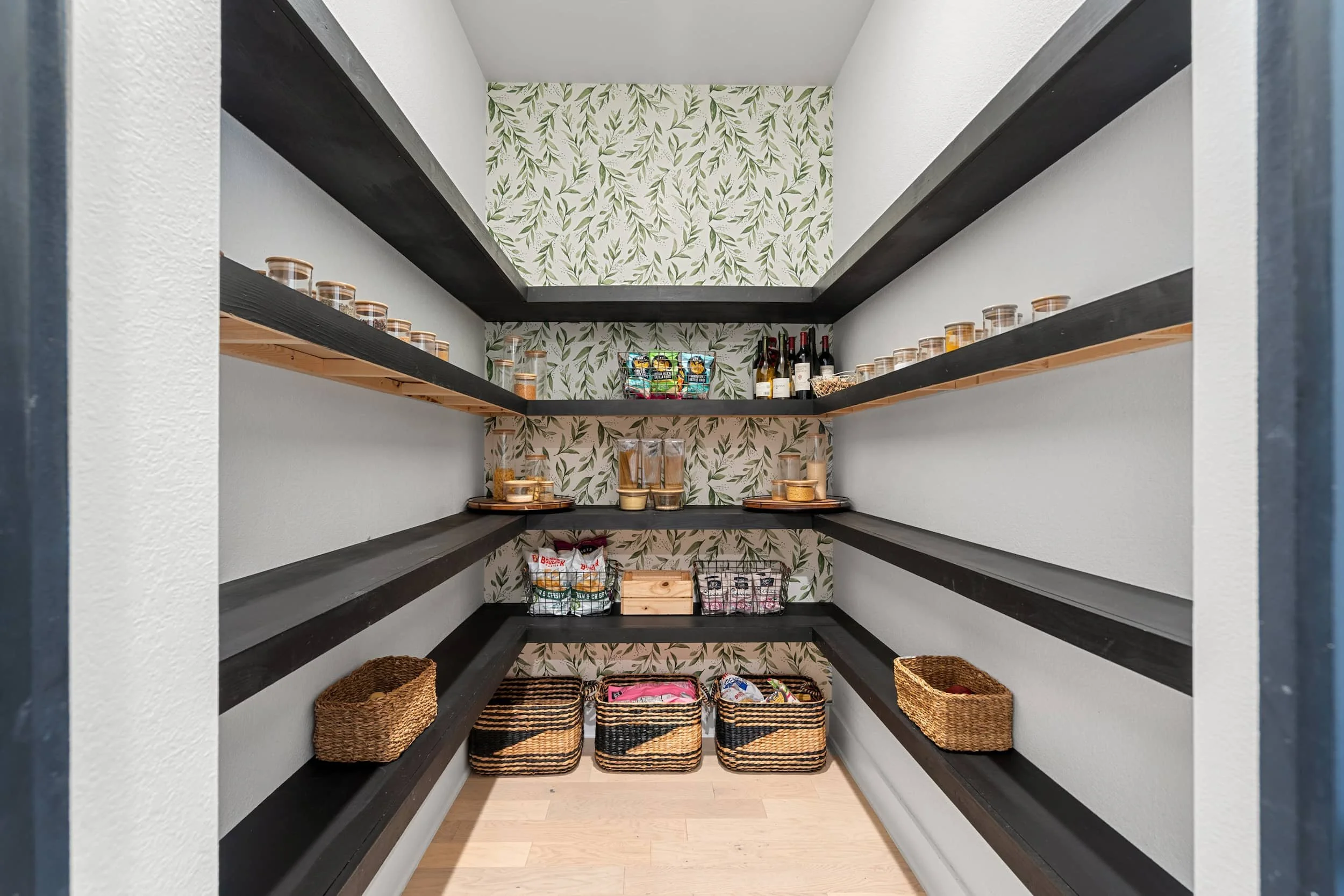 Pantry with open shelving, black wood shelves, storage baskets, organized layout, patterned wallpaper.