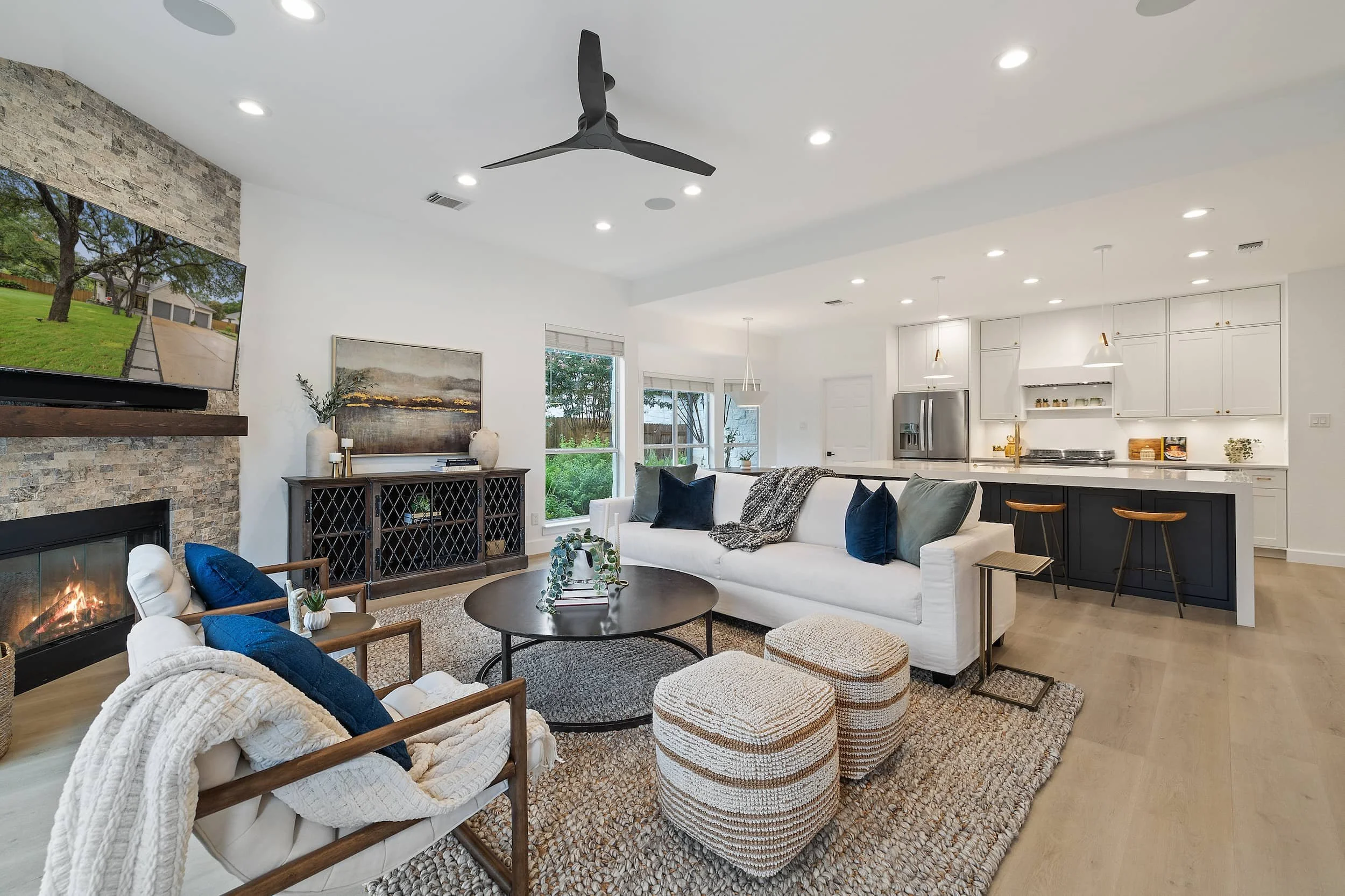 Bright open living room with stone fireplace and TV, white sofa and chairs, round coffee table, and a modern kitchen with island and bar seating.