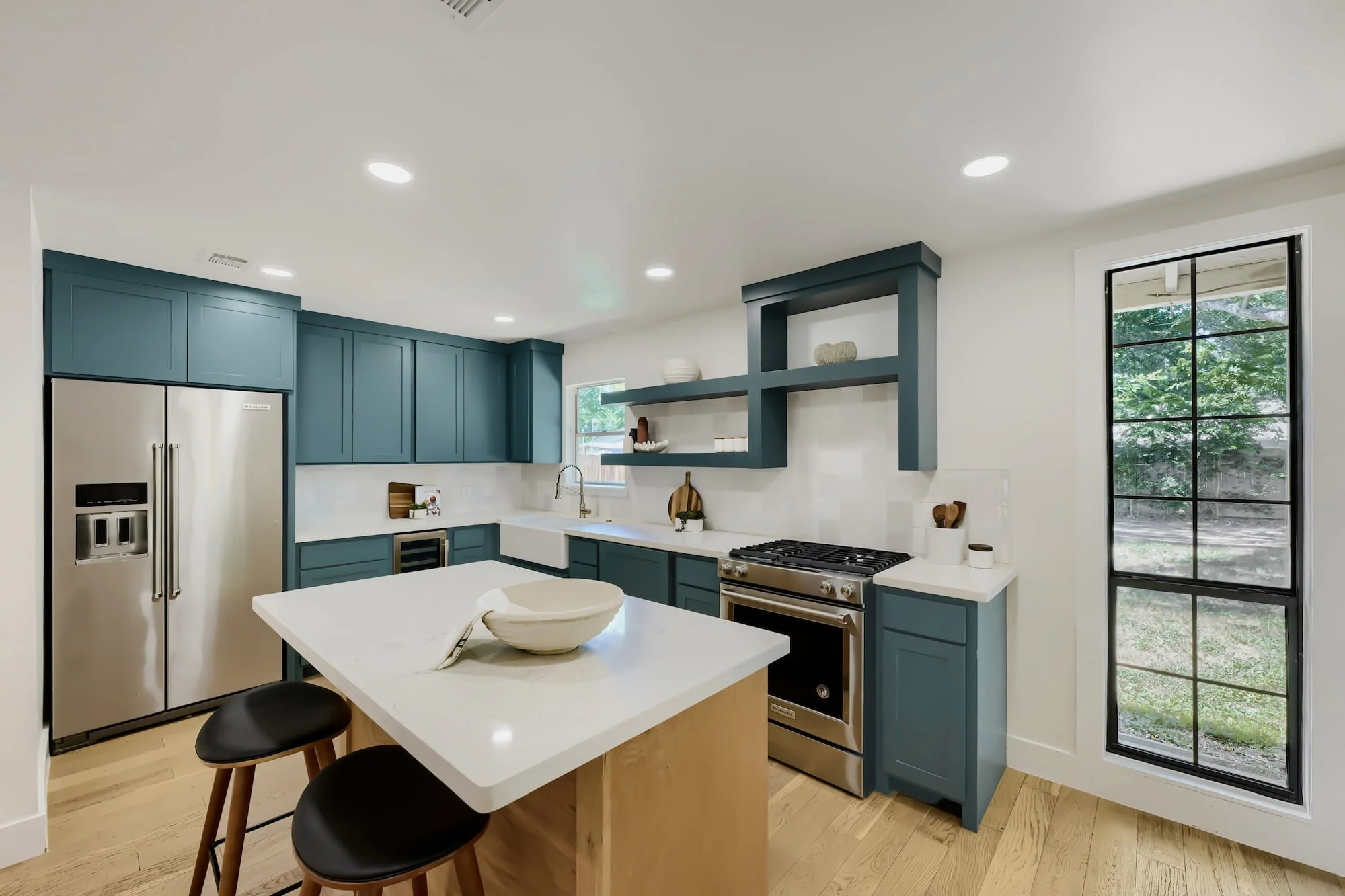 Staged kitchen with teal cabinets, a light wood island with black leather stools, and a tall steel-framed window.