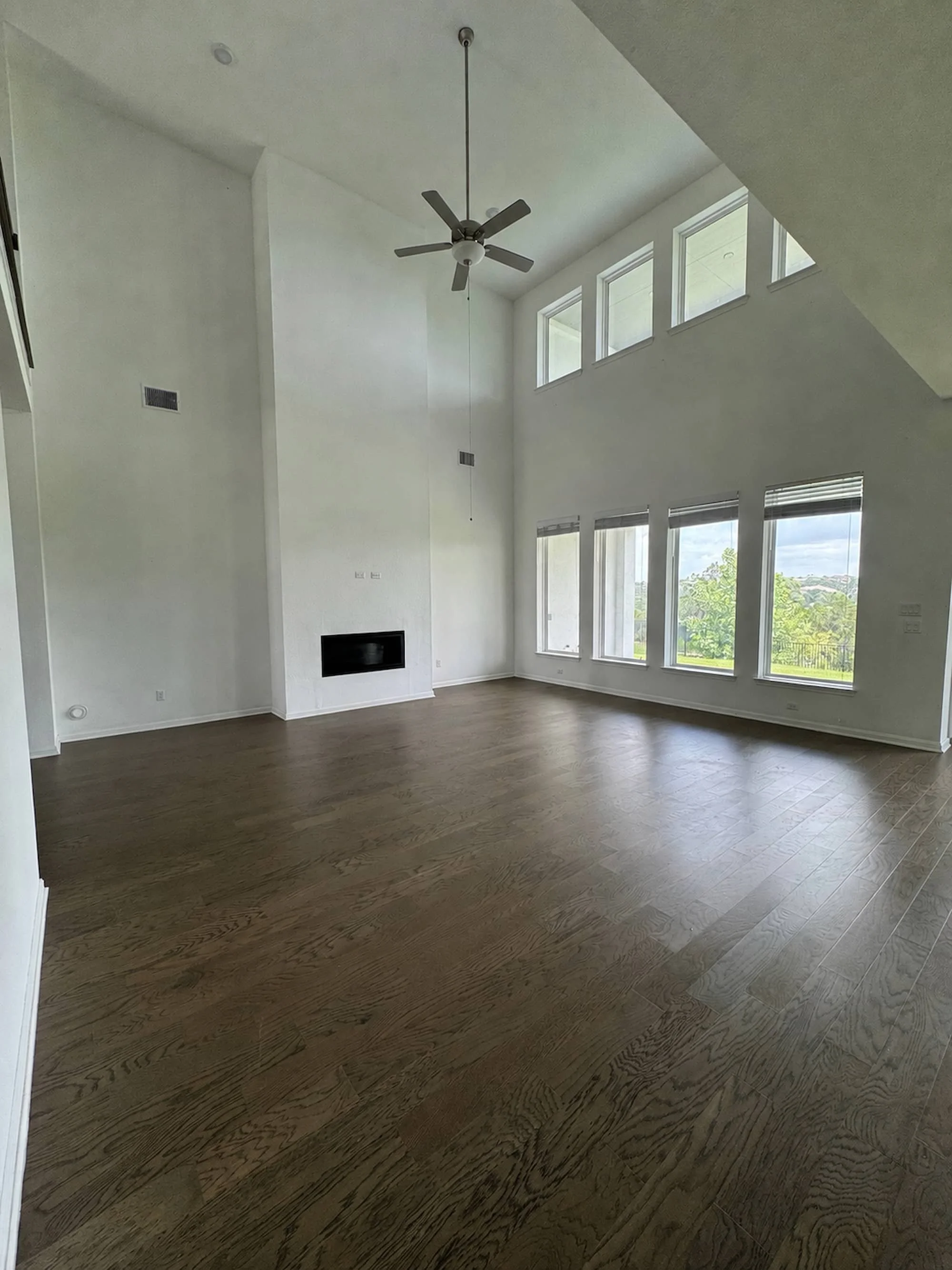 Vacant great room with soaring vaulted ceilings, a ceiling fan, a wall of tall windows with tree views, dark hardwood flooring, and a modern linear fireplace built into the wall.