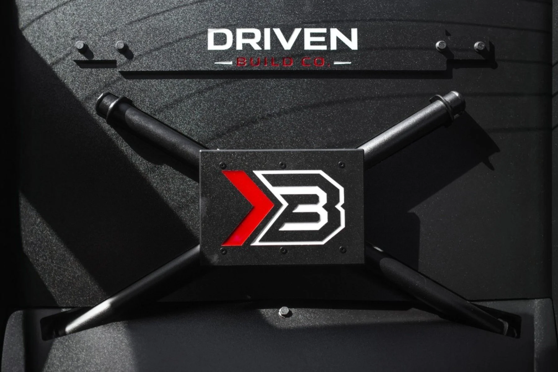Close-up of a black surface with a logo featuring a red and white arrow pointing right and the letter 'B', with a rectangular metal frame behind it. Above, there is a sign that reads 'DRIVEN BUILD CO.' in white and red letters.