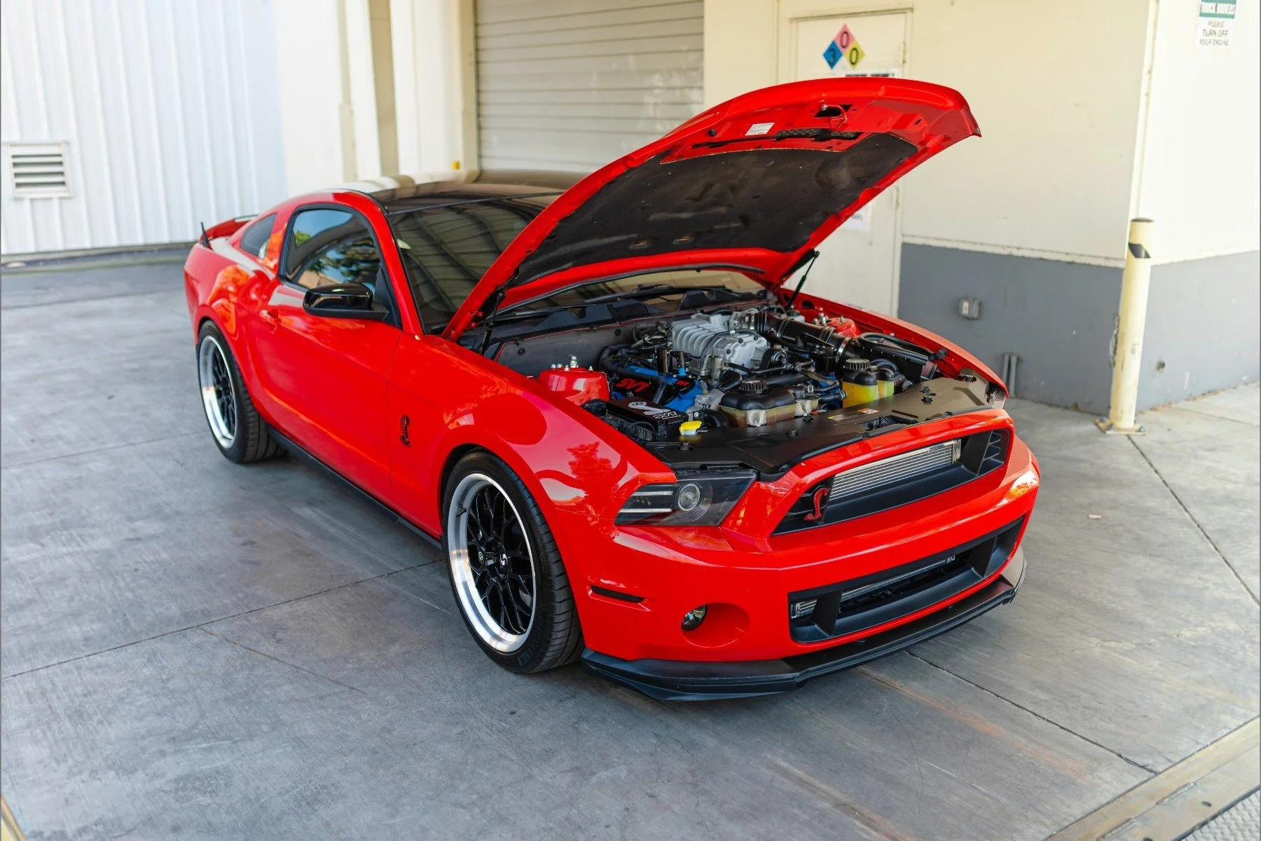 A red sports car with its hood open revealing the engine, parked in an industrial or garage area.