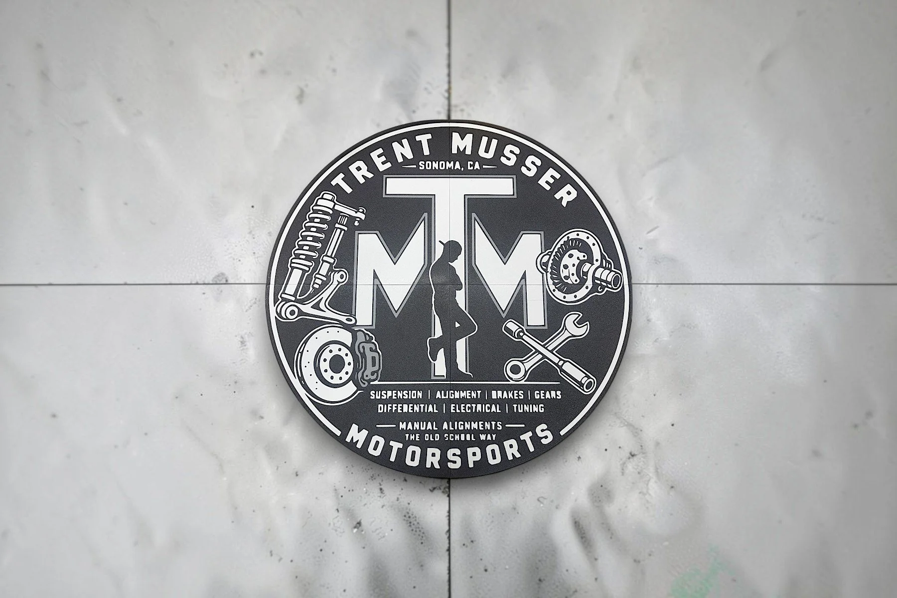 A circular black and white sign for Turrent Musser Motorsports in Sonoma, CA. The sign features a silhouette of a person leaning against a large letter T, with illustrations of car suspension, brake disc, gear, and wrench. The text lists services lik