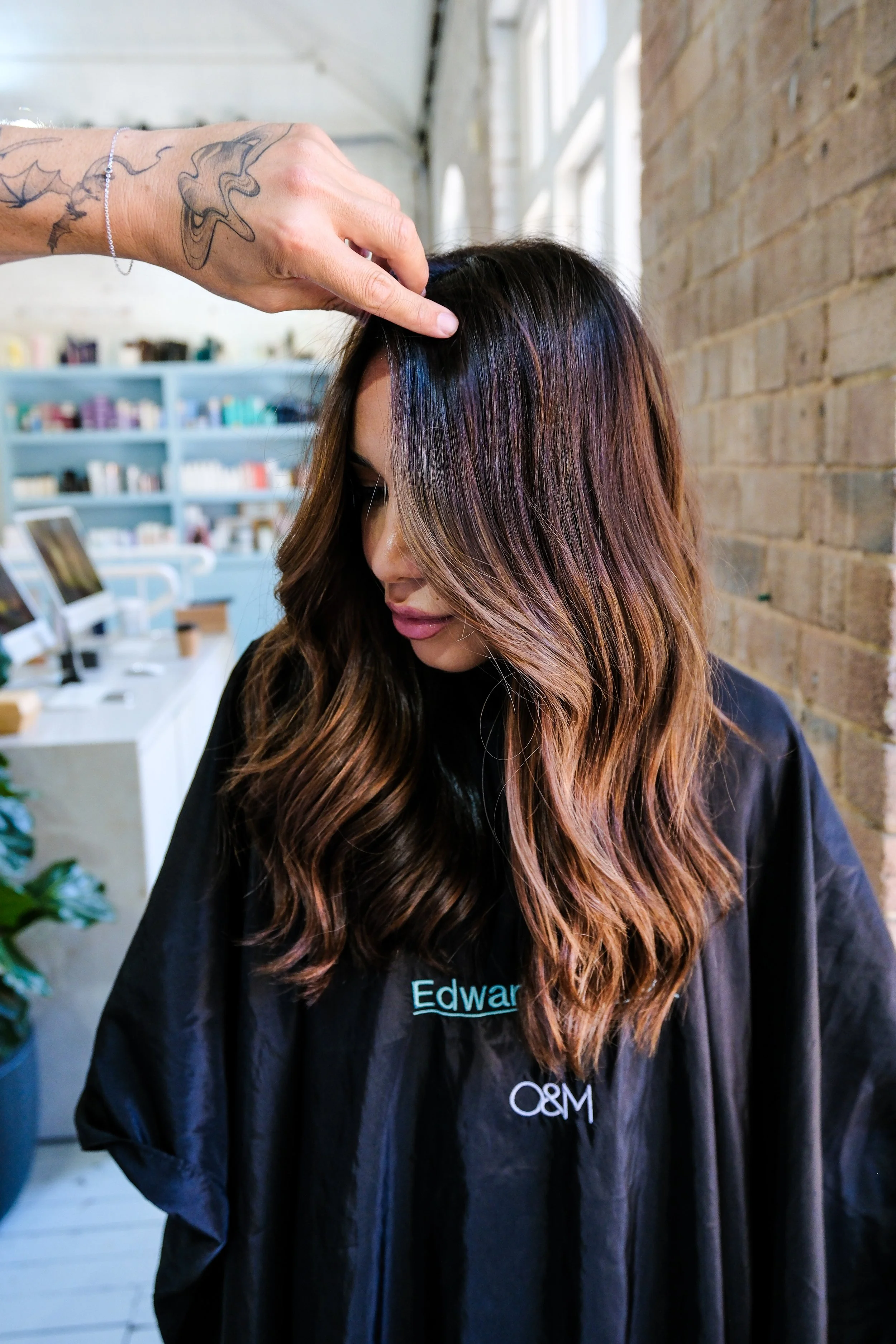A woman in a black salon cape with turquoise embroidered name tags, sitting in a hair salon, while a stylist touches her hair. The salon has a brick wall and shelves with hair products in the background.