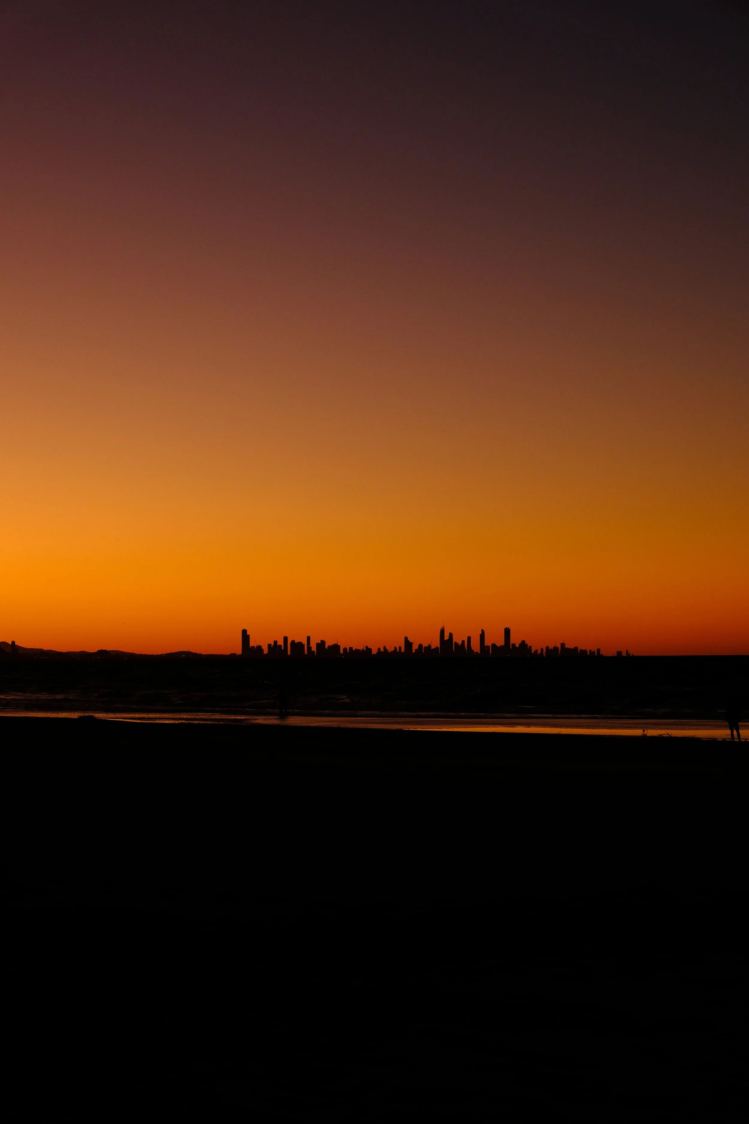 Sunset over a city skyline with dark silhouetted buildings, visible towards the horizon, and a calm waterfront in the foreground.