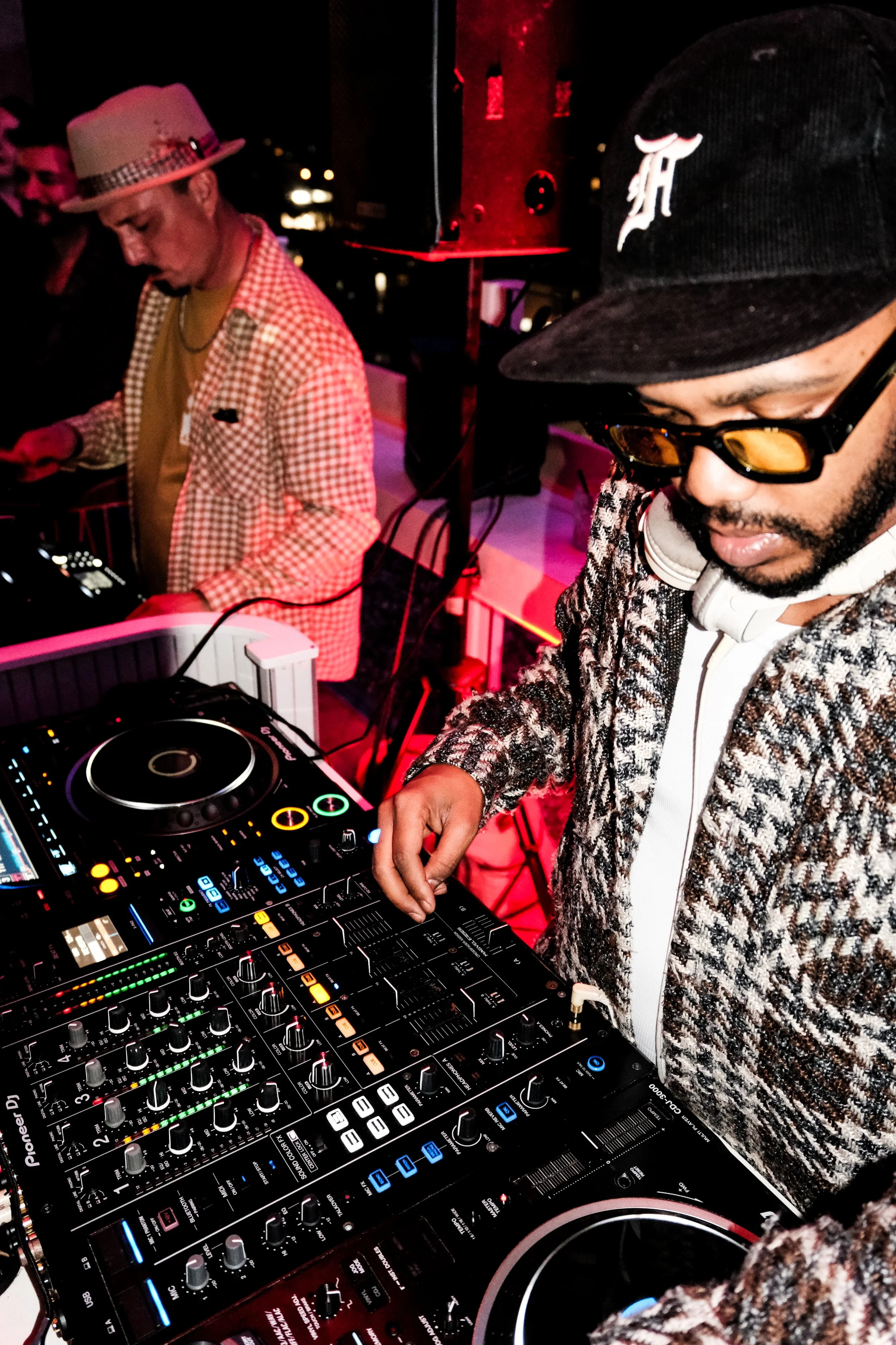 Two DJs performing at a night event, one in a patterned blazer and cap with headphones around his neck, and another in a checkered jacket and tan hat, focused on their DJ equipment with colorful lights.