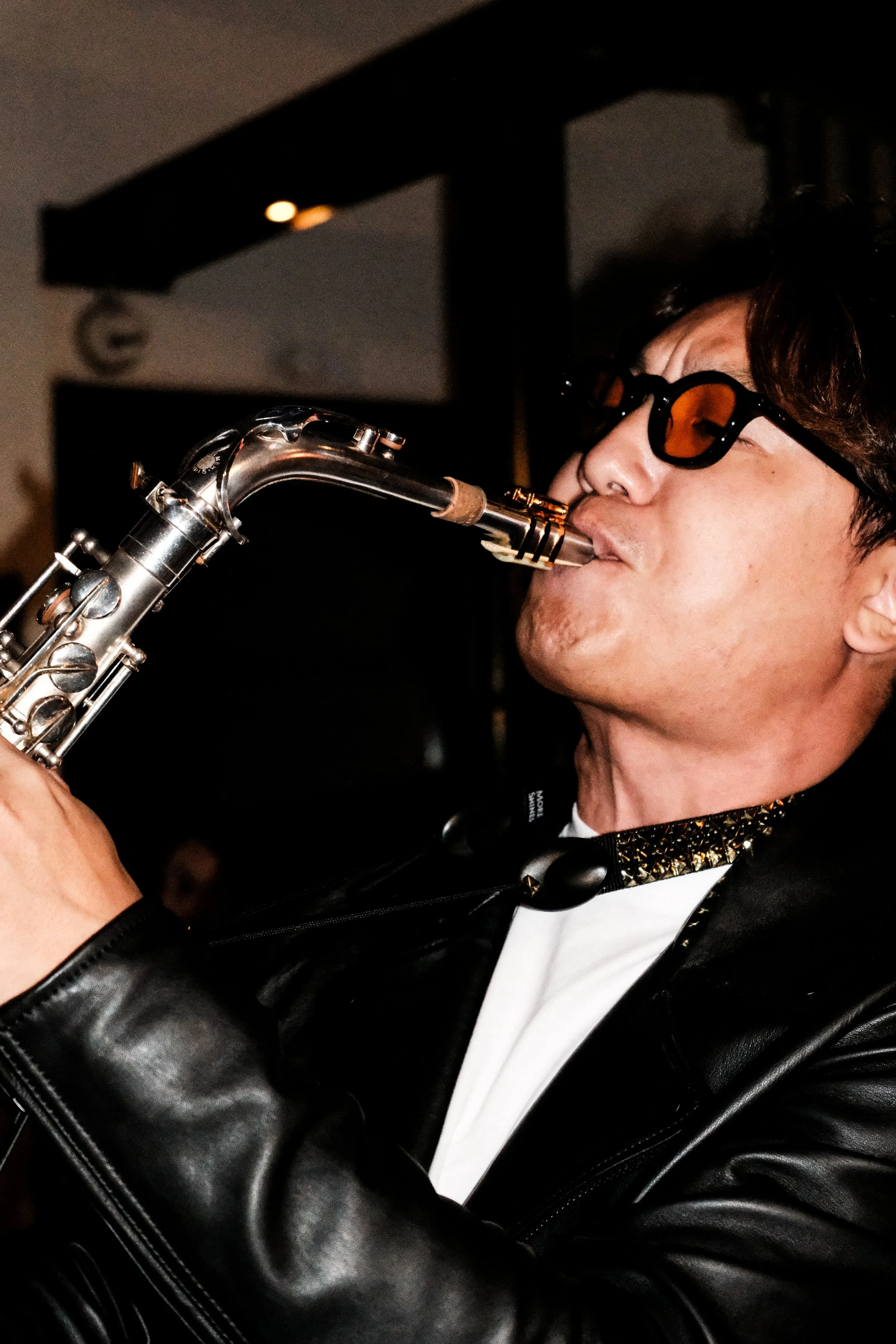 A man wearing sunglasses and a leather jacket playing a saxophone indoors.