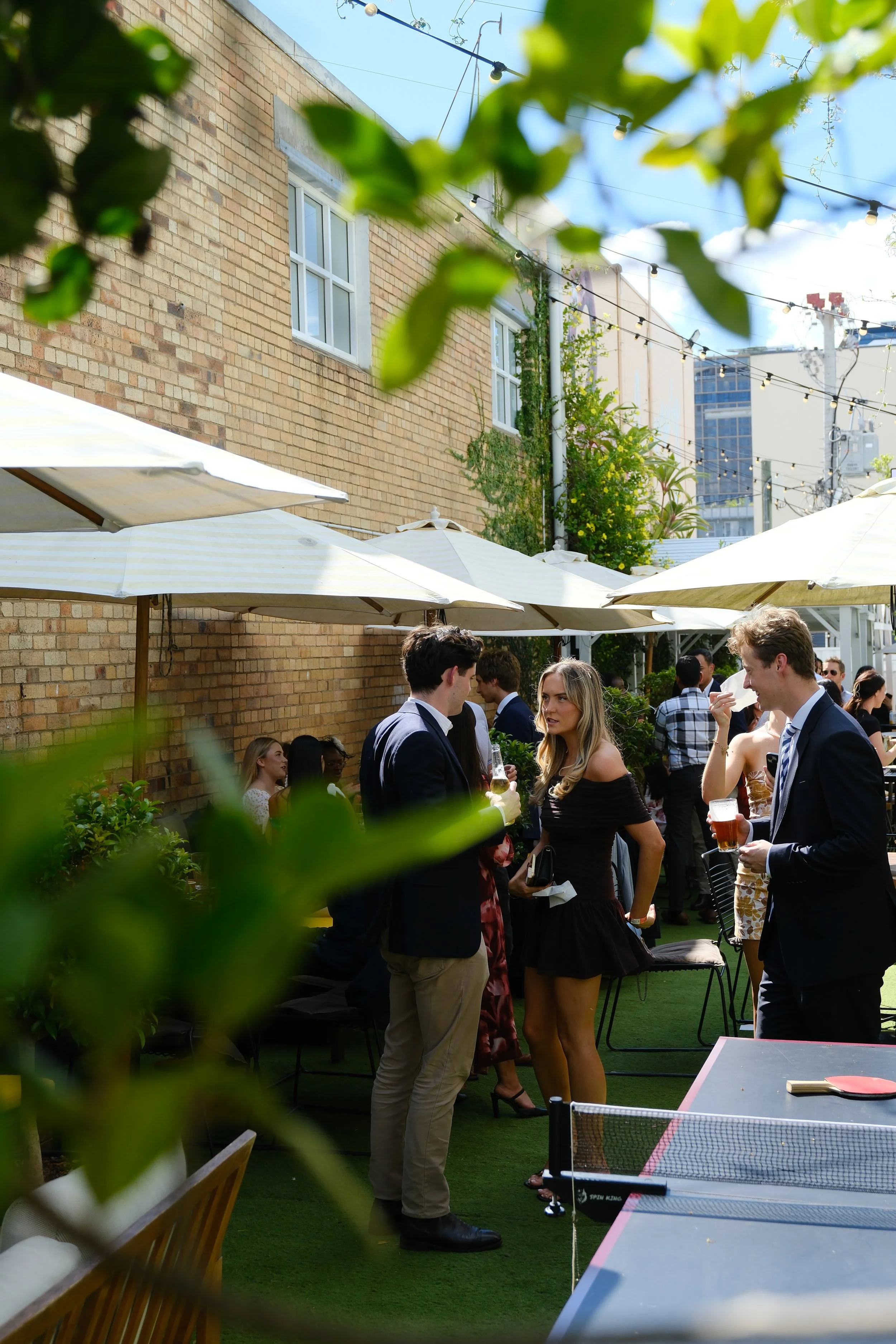 People socializing outdoors at a patio party with umbrellas, a brick building, and string lights overhead.