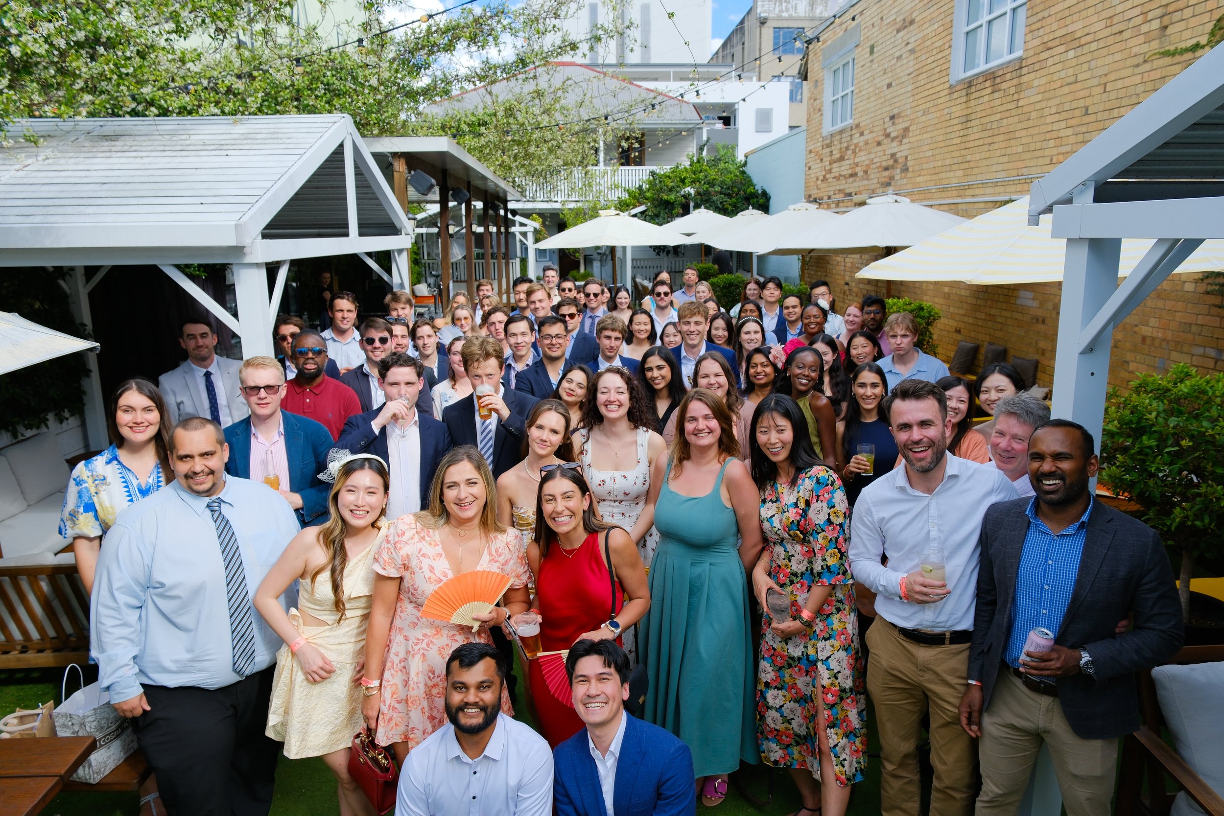 A large group of diverse people gathered at an outdoor party or celebration, smiling and holding drinks, with wooden structures and umbrellas in the background.