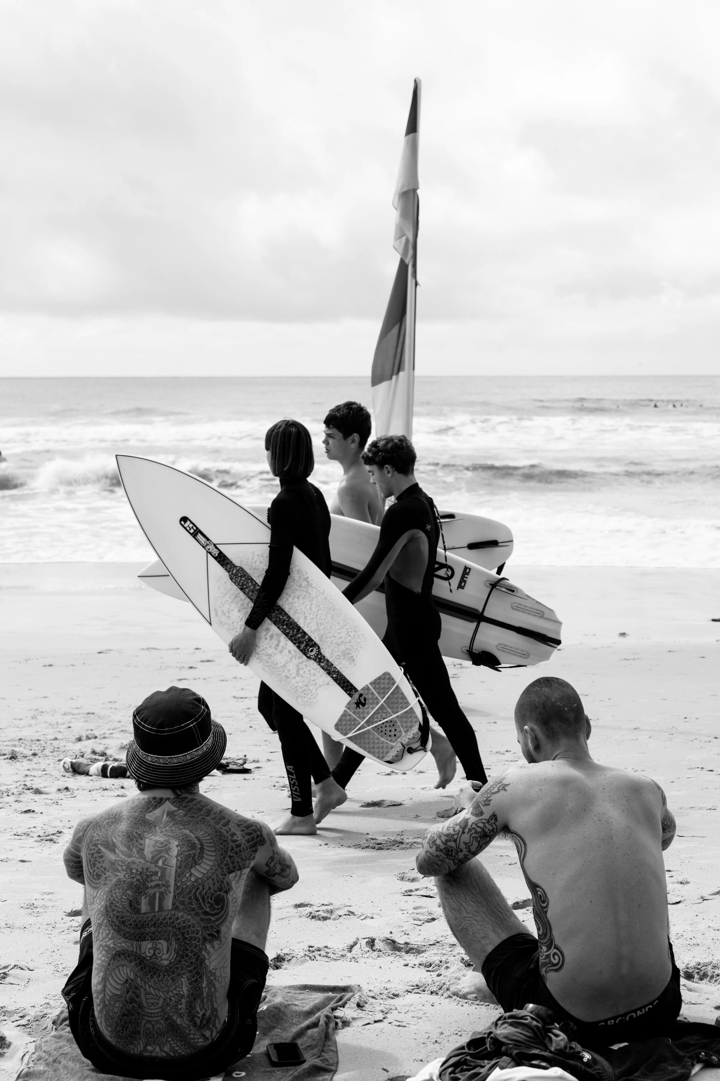 People with surfboards walking on the beach, with two tattooed men sitting on the sand in the foreground and weathered clouds in the sky.