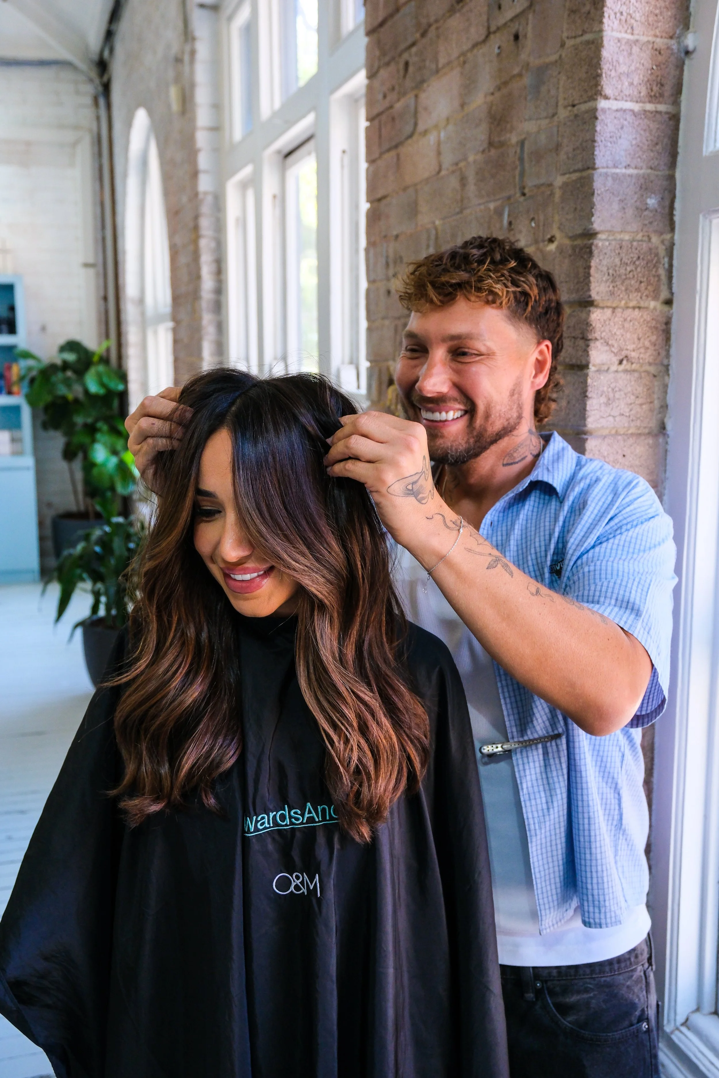 A hairstylist smiling as he trims a woman's hair inside a bright salon with large windows and exposed brick walls.