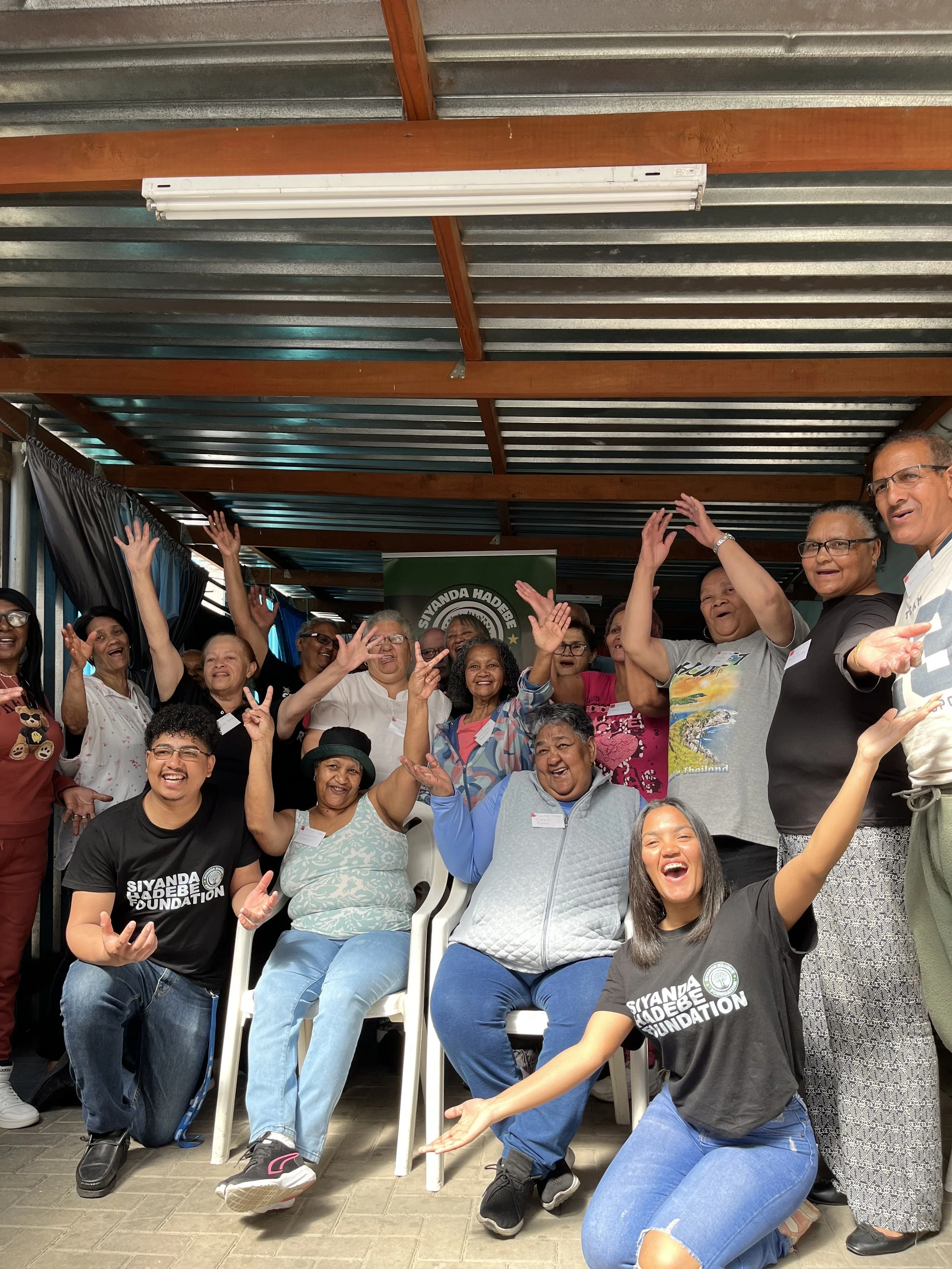 Group of diverse people smiling and raising hands, posing for a photo indoors, some wearing Siyanda Hadebe Foundation T-shirts.