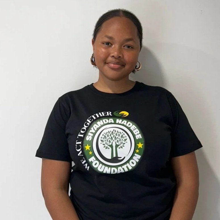 A woman wearing a black T-shirt with the logo of the Siyanda Hadebe Foundation, standing against a plain white wall.