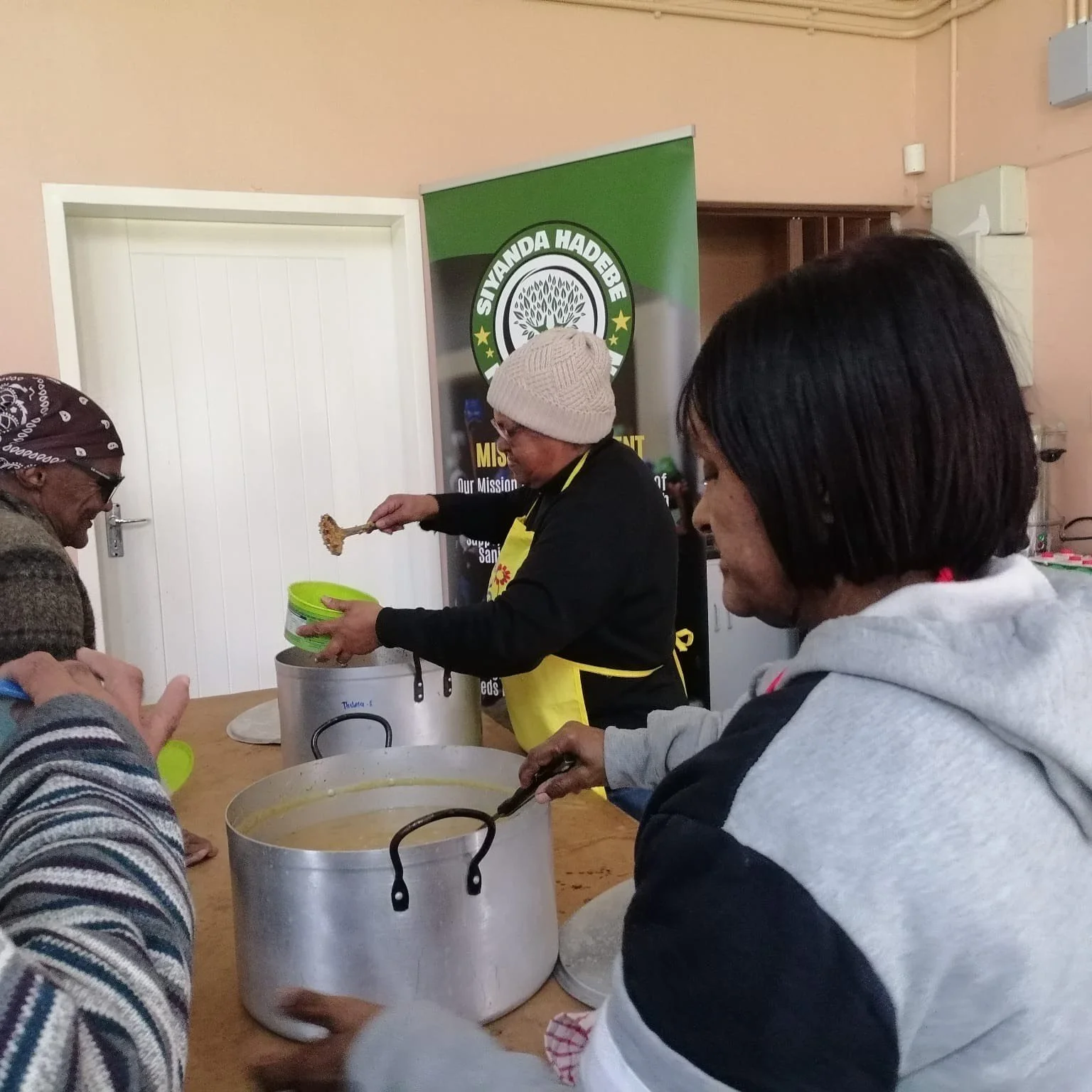 People serving food in a community kitchen, with a woman in a beanie ladling soup from a large pot. There are other individuals waiting, and a banner with the words 'Siyanda Hadebe Foundation' in the background.