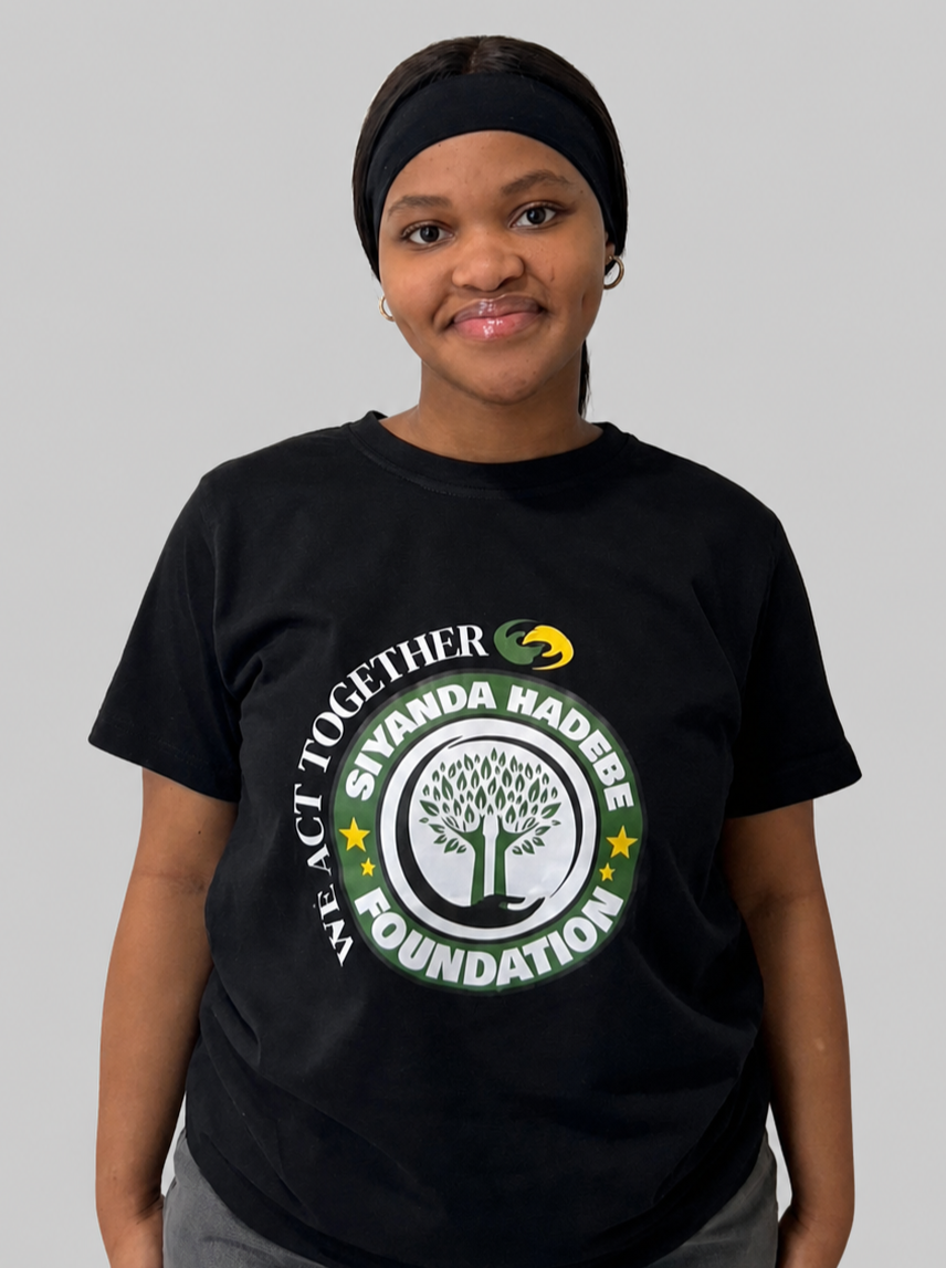 A woman wearing a black T-shirt with a logo that says 'We Act Together' and 'Siyanda Hadebe Foundation,' standing against a plain white wall.