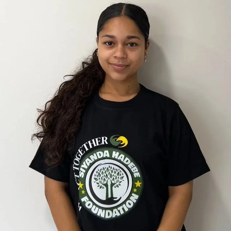 Young woman with dark, curly hair in a ponytail, wearing a black T-shirt with a logo that says 'We Act Together Siyanda Hadebe Foundation' and an image of a tree, standing against a plain white wall.