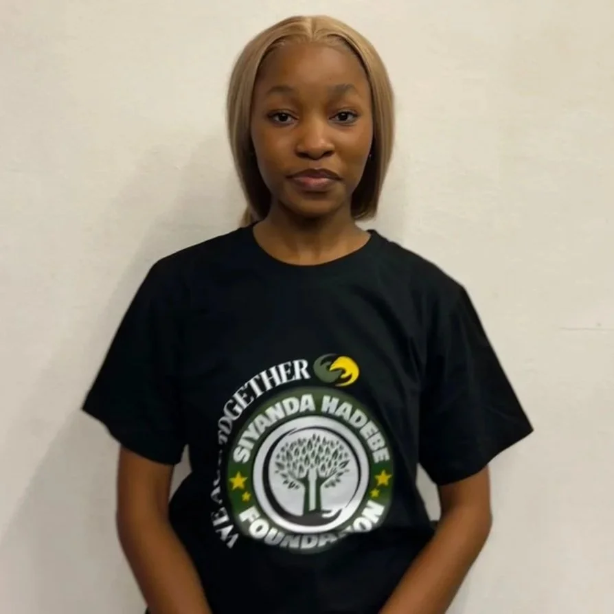 A young woman with blonde hair standing against a plain white wall, wearing a black T-shirt with a logo that says 'We Act Together' and 'Siyanda Hadebe Foundation,' standing against a plain white wall.