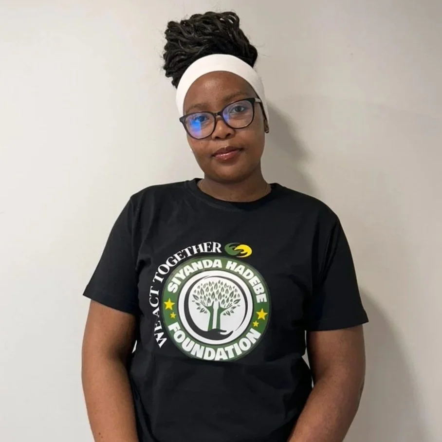 A woman wearing glasses, a white headband, and a black t-shirt with a logo that reads 'We Act Together - Siyanda Hadebe Foundation,' standing against a plain wall.