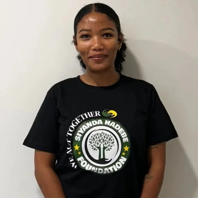 A young woman with a smile, wearing a black T-shirt that reads "Siyanda Hadebe Foundation" with a tree logo and text around it, standing against a plain white wall.