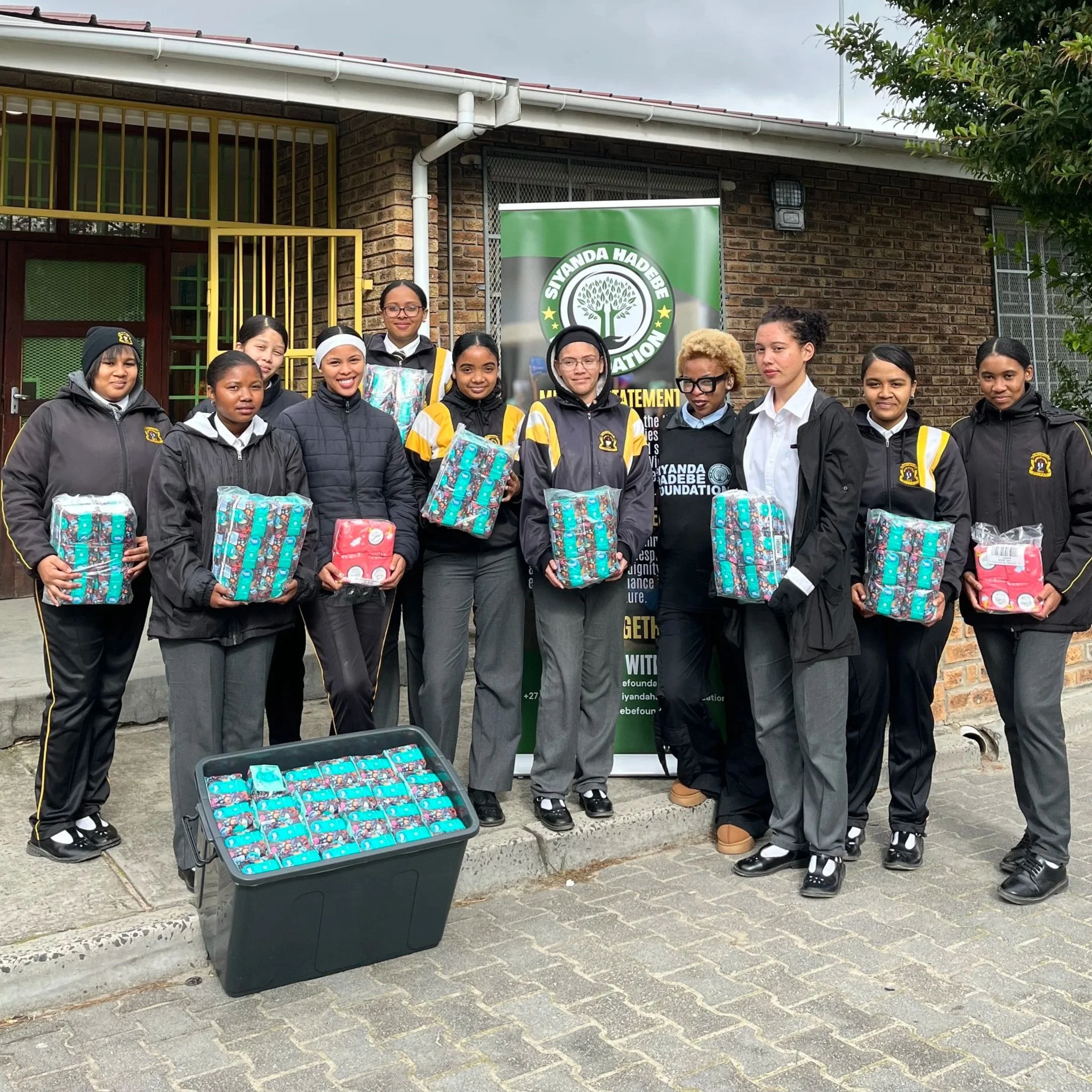 Group of girls standing outdoors, holding sanitary pads, in front of a banner for the Siyanda Hadebe Foundation.
