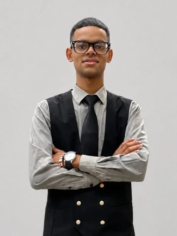 A young man with glasses wearing a black t-shirt with a logo that says 'We Act Together' and 'Siyanda Hadebe Foundation,' standing against a plain white wall.