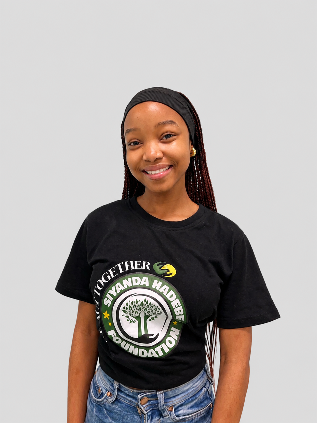 A woman wearing glasses, a white headband, and a black t-shirt with a logo that reads 'We Act Together - Siyanda Hadebe Foundation,' standing against a plain wall.
