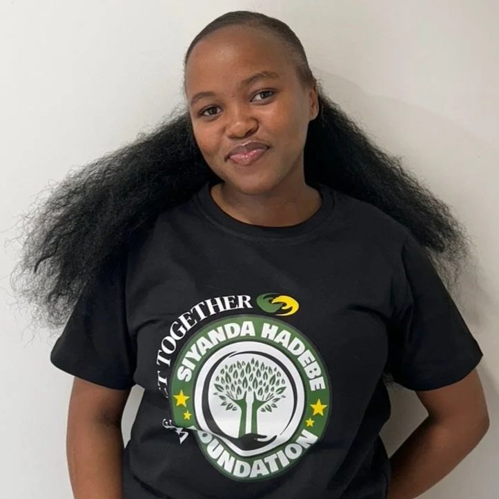 Young woman with curly hair wearing a black T-shirt with a logo that says 'We Act Together' and 'Siyanda Hadebe Foundation,' standing against a plain white wall.