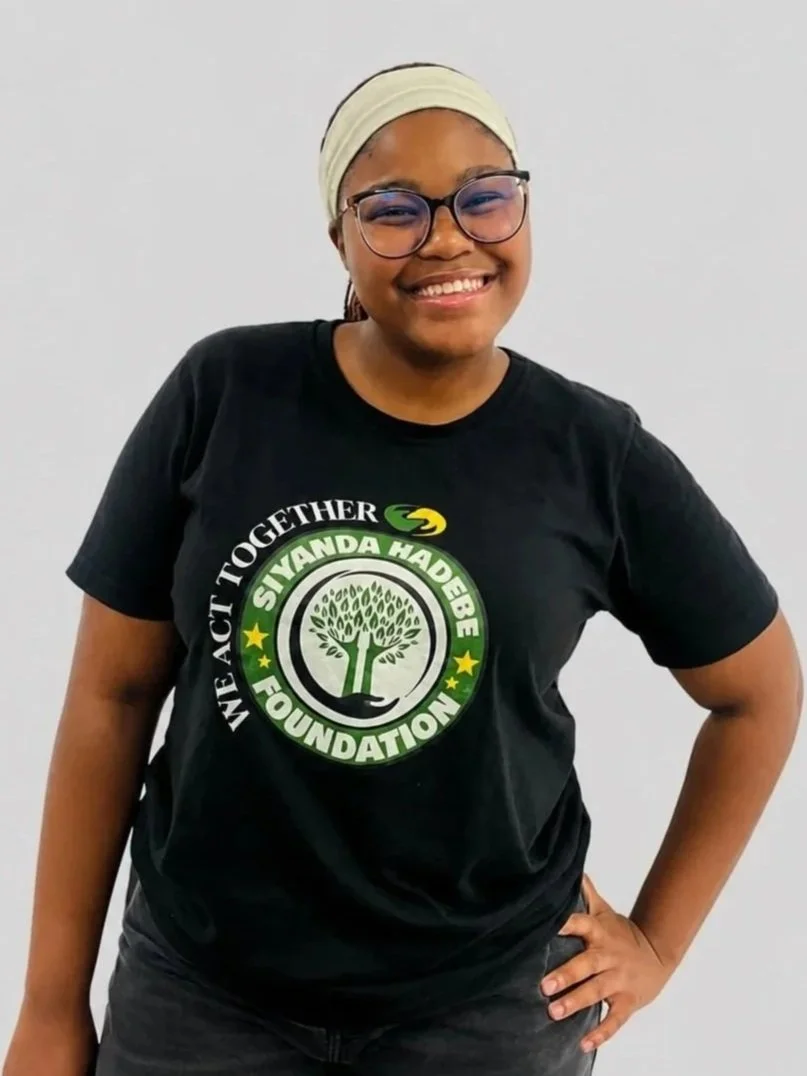 Smiling young woman wearing a black T-shirt with a logo for the Siyanda Hadebe Foundation, standing against a plain white wall.