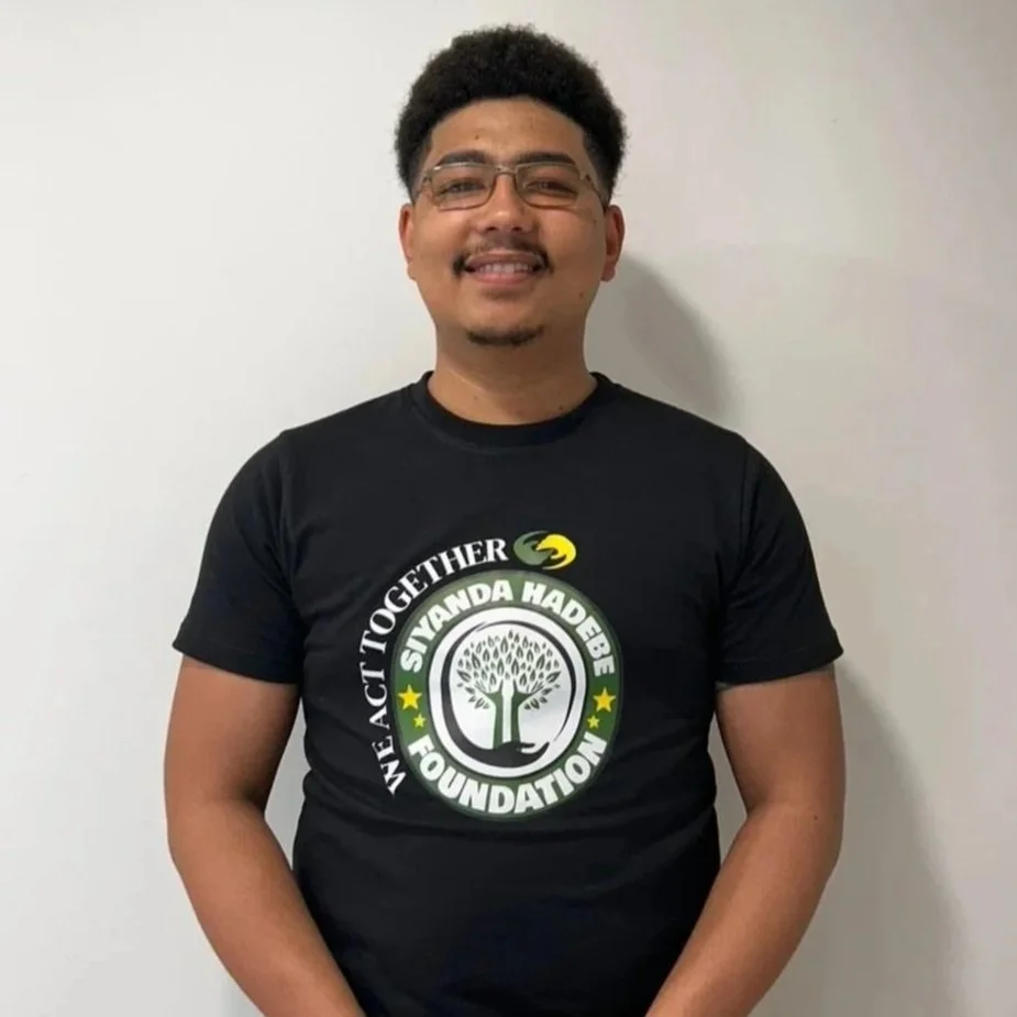A smiling man with glasses and a beard, wearing a black T-shirt with a logo that says 'We Act Together' and 'Siyanda Hadebe Foundation,' standing against a plain white wall.