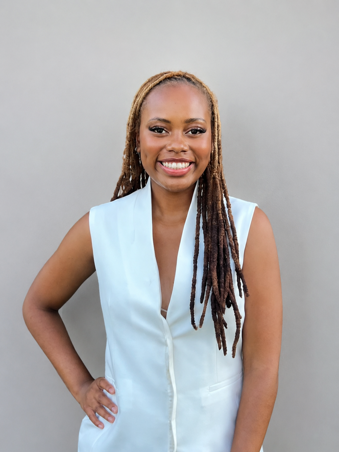 A woman with glasses and hoop earrings wearing a white headband, a white shirt, and a navy blazer, standing against a plain light gray background.
