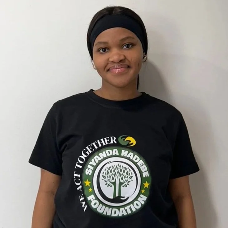 A woman wearing a black T-shirt with a logo that says 'We Act Together' and 'Siyanda Hadebe Foundation,' standing against a plain white wall.