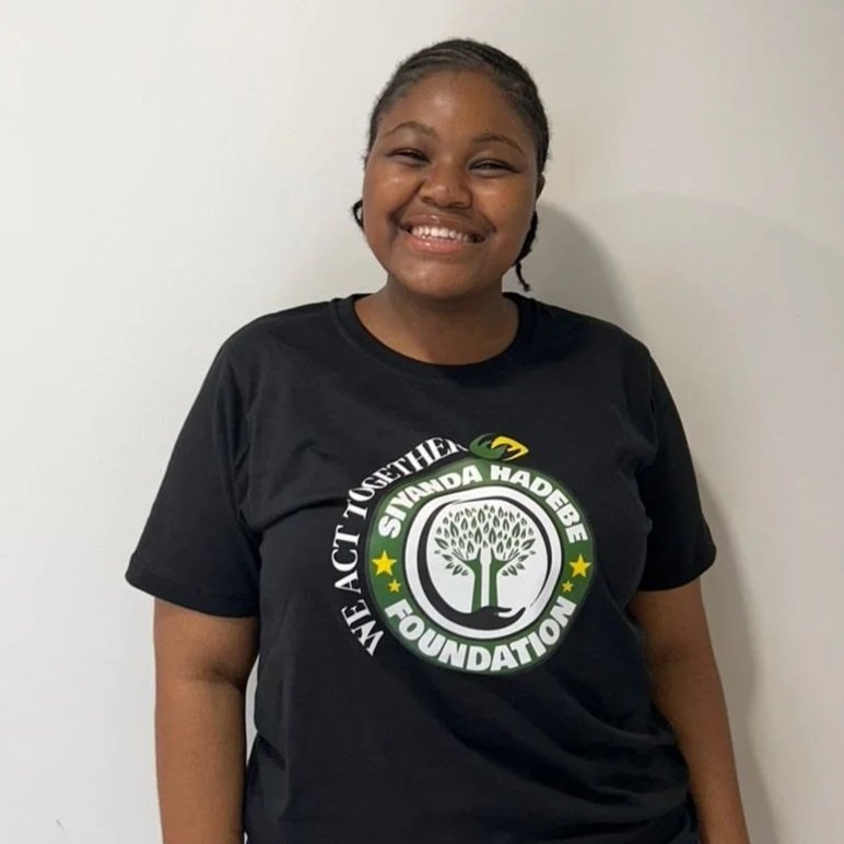Smiling young woman wearing a black T-shirt with a logo for the Siyanda Hadebe Foundation, standing against a plain white wall.