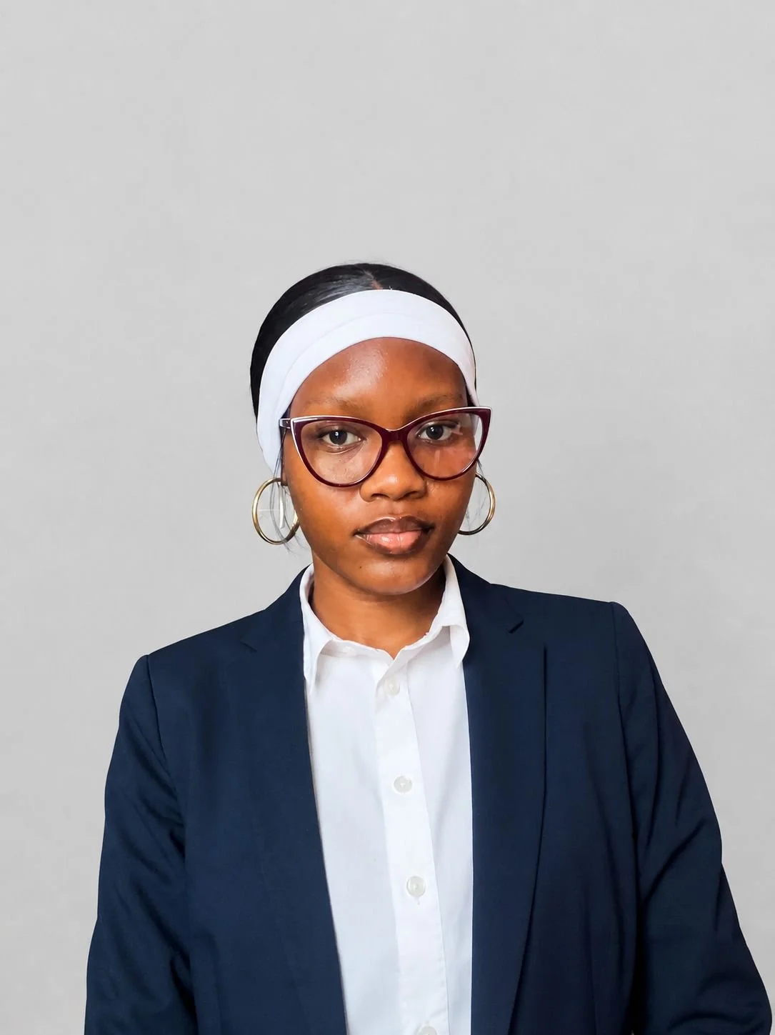 A woman with glasses and hoop earrings wearing a white headband, a white shirt, and a navy blazer, standing against a plain light gray background.