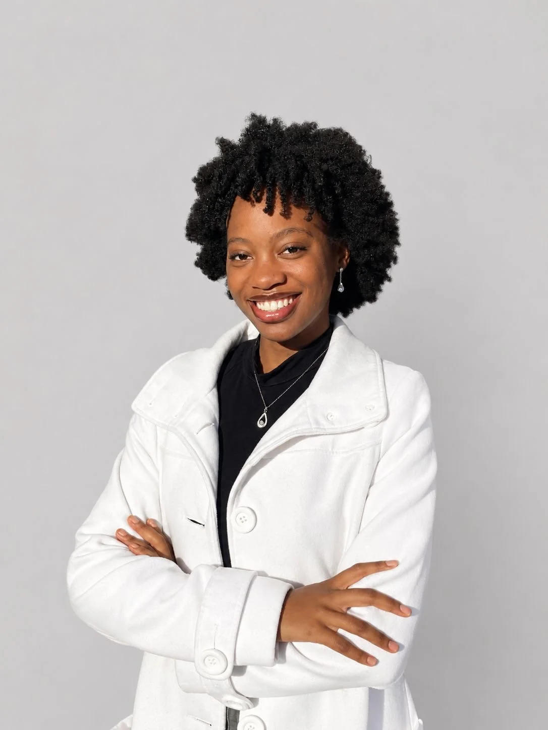 A young Black woman with a natural curly hairstyle, wearing a white jacket, black top, and jewelry, smiling with arms crossed against a plain light gray background.