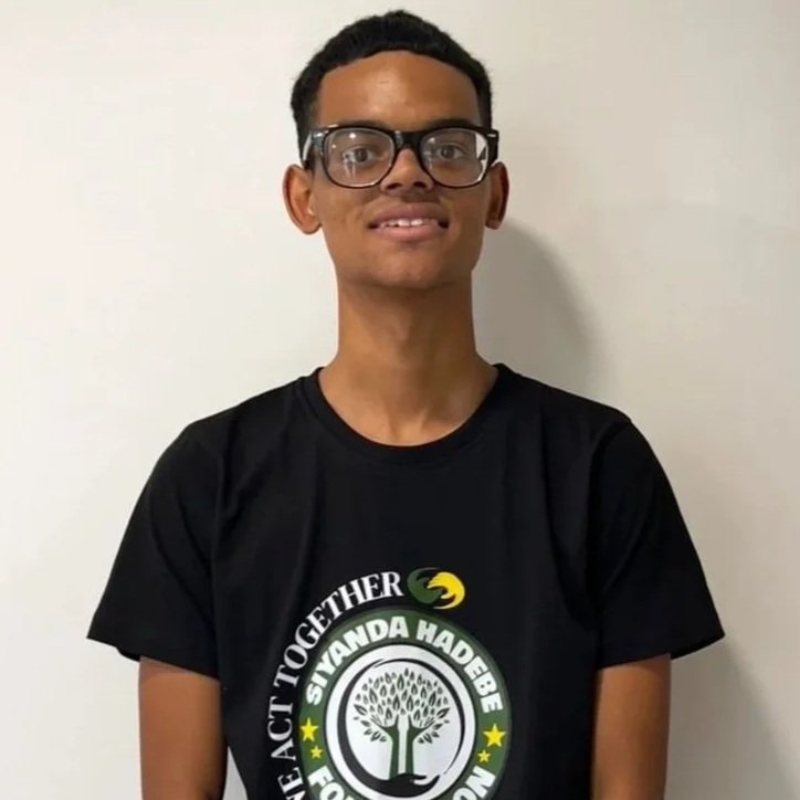 A young man with glasses wearing a black t-shirt with a logo that says 'We Act Together' and 'Siyanda Hadebe Foundation,' standing against a plain white wall.