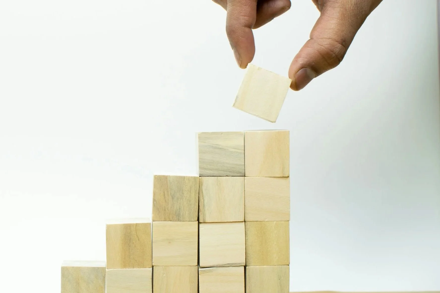 A person's hand is placing a wooden block on top of a pyramid of wooden blocks, constructing a staircase-like structure against a plain, white background.