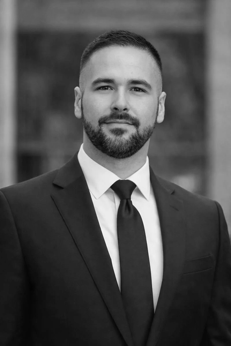 Black and white portrait of a man in a suit with a tie, smiling slightly, with a blurred urban background.
