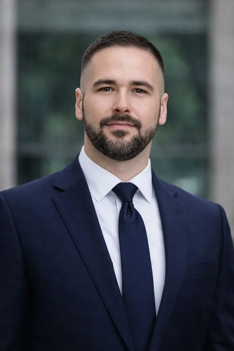 A man in a navy suit and tie, with short dark hair and a beard, standing outdoors with a modern glass building in the background.