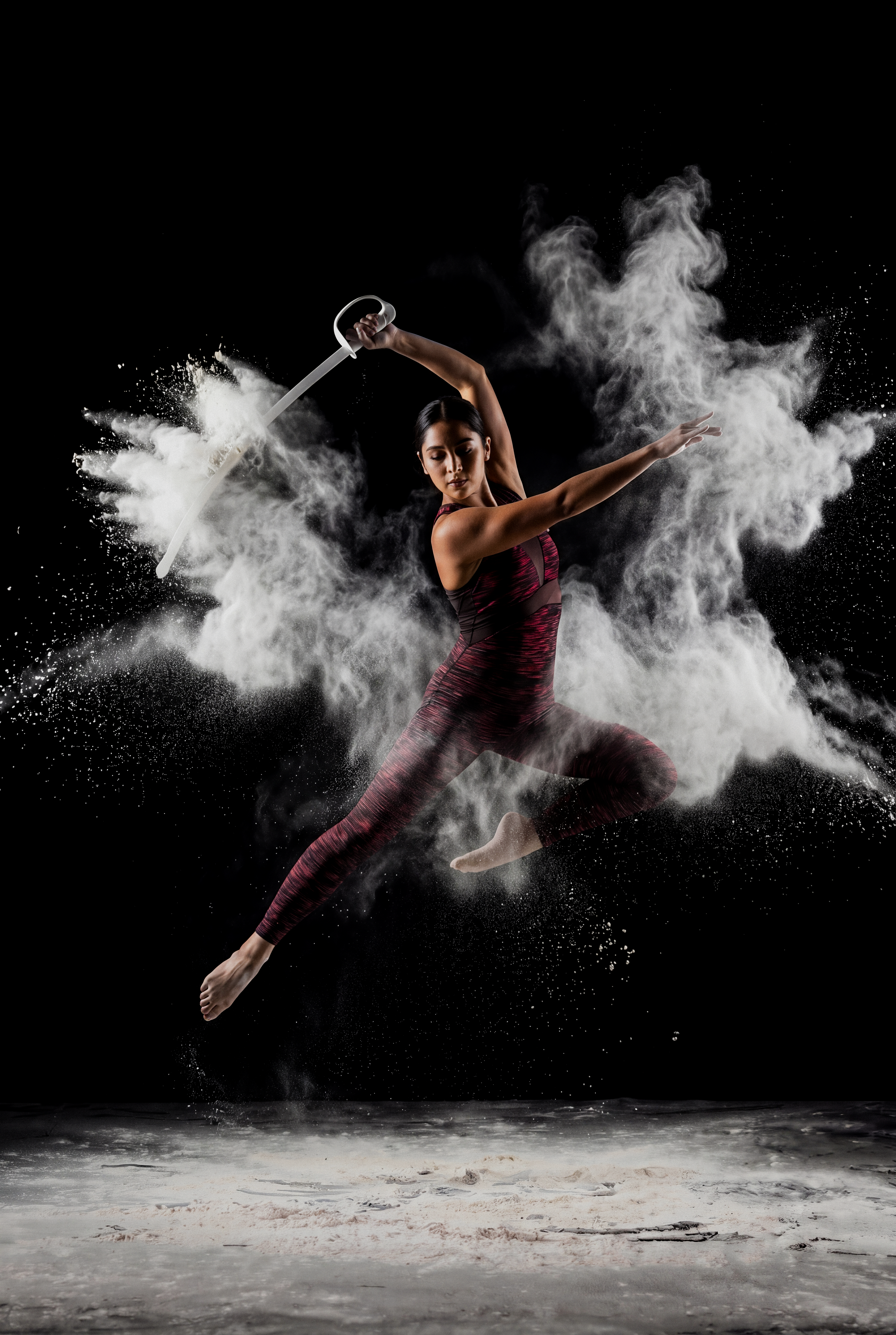A woman in athletic clothing performing a dance move with dust or powder exploding around her, against a black background.