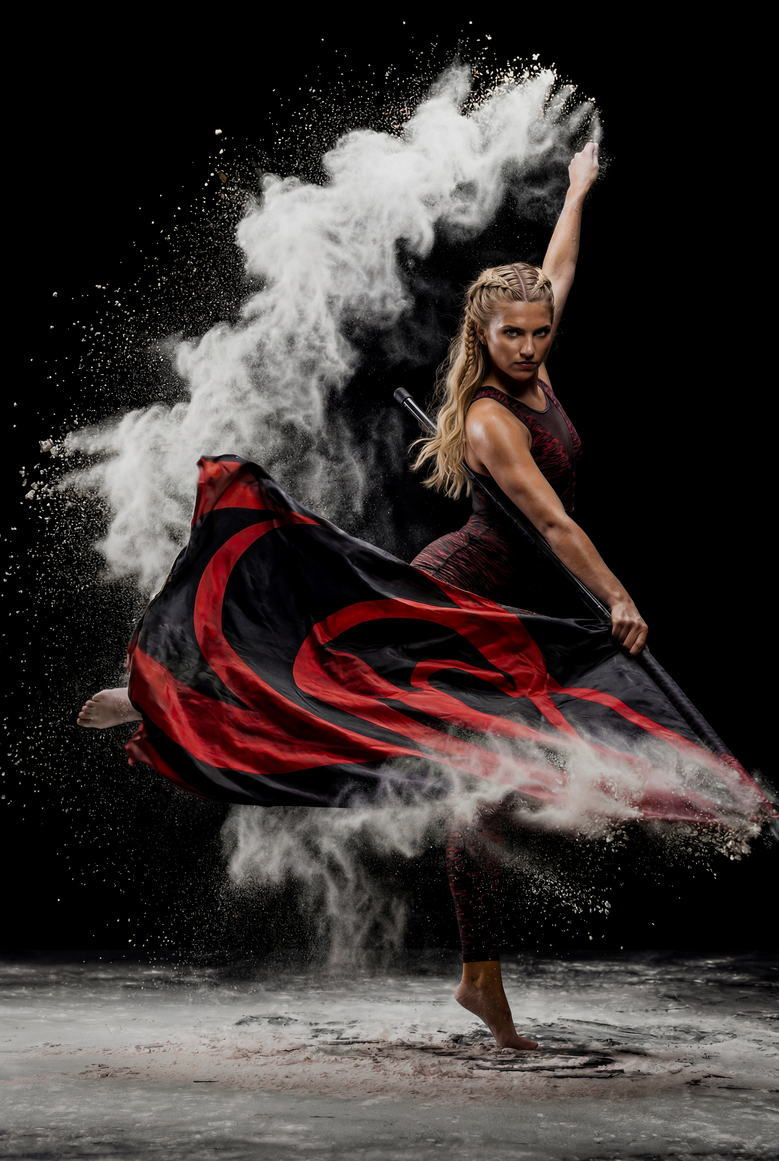 A woman performing a dance move on a sandy surface with black background, holding a black and red flag and surrounded by white powder or dust.