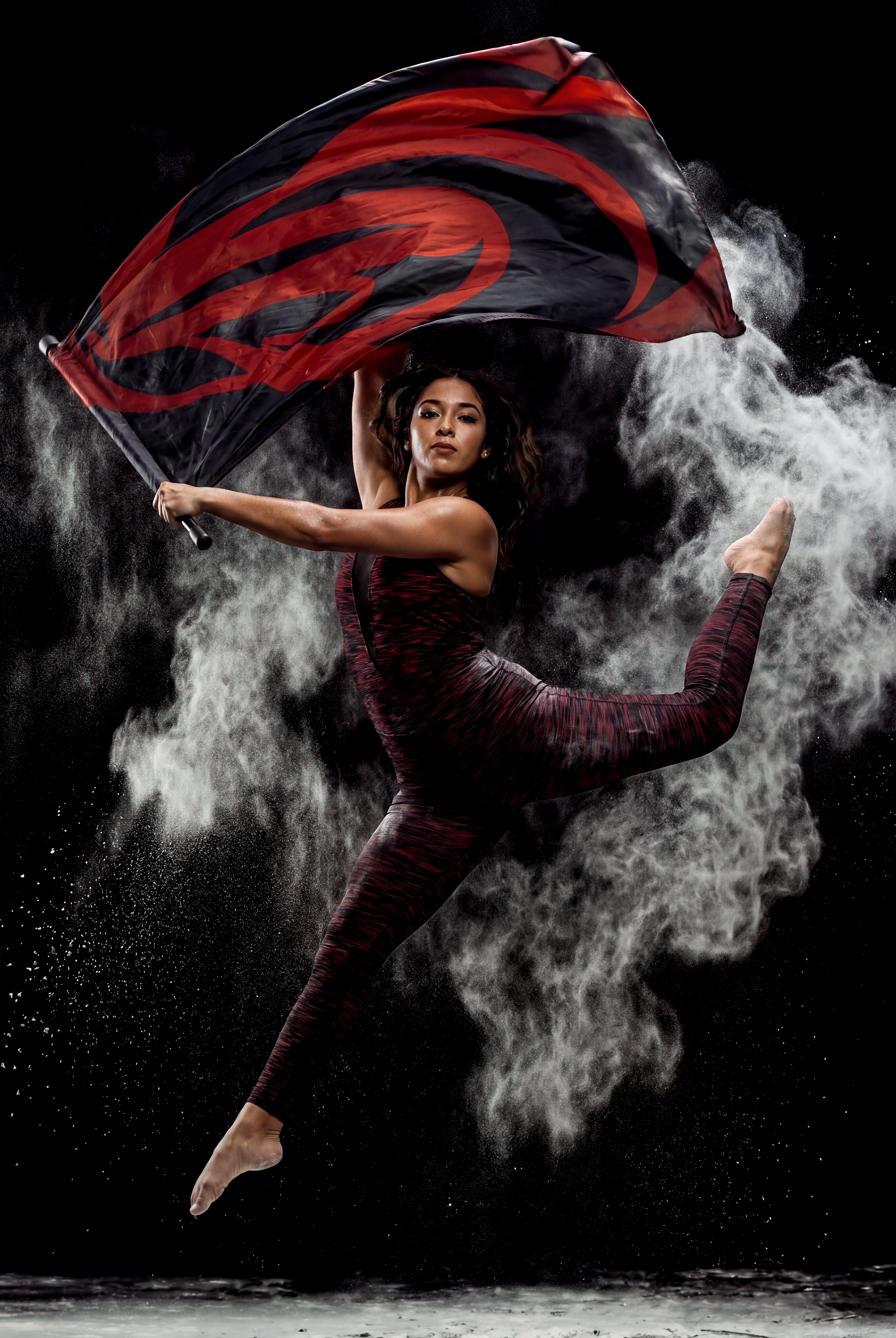 A woman performing a dance or athletic move, holding a black and red flag, with water spray and a dark background.