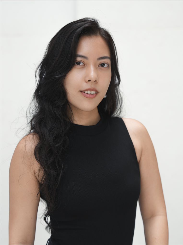 A young woman with long wavy black hair and light skin, wearing a black sleeveless top, posing against a white background.