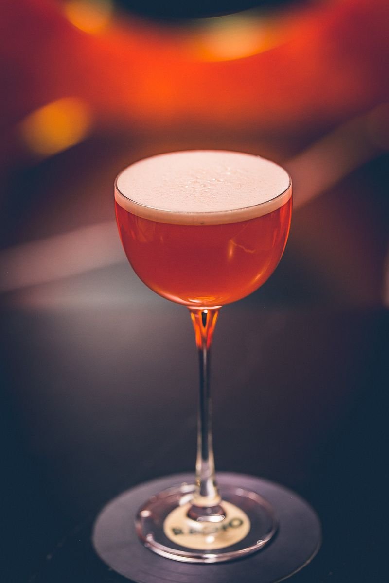 A close-up of a cocktail glass filled with a reddish-orange drink topped with a frothy foam, set against a dark background with blurred colorful lights.