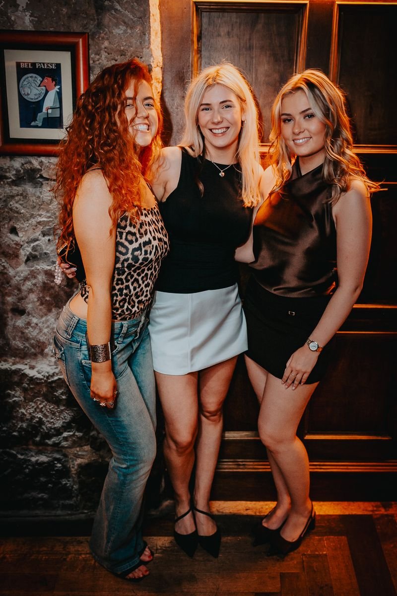 Three young women smiling and posing together inside a dimly lit venue with stone walls and framed artwork in the background.