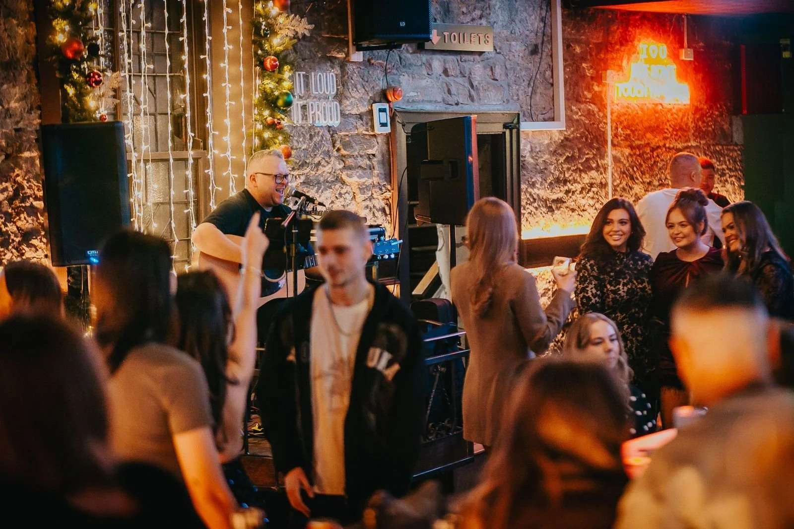 People dancing and socializing in a dimly lit bar or club decorated with Christmas ornaments and string lights, with a musician playing guitar on stage.