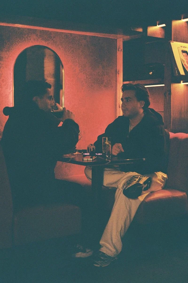 Two young men sitting at a table in a dimly lit bar or restaurant, engaging in conversation. One is drinking from a mug, and the other is sitting with his hands folded. There is a drink with a straw and a bottle on the table, and a TV screen mounted on the wall in the background.