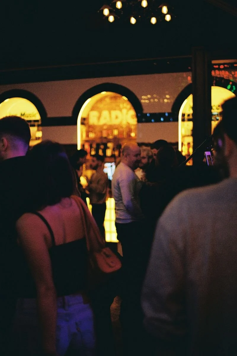 People gathered in a dimly lit bar with illuminated signage in the background.