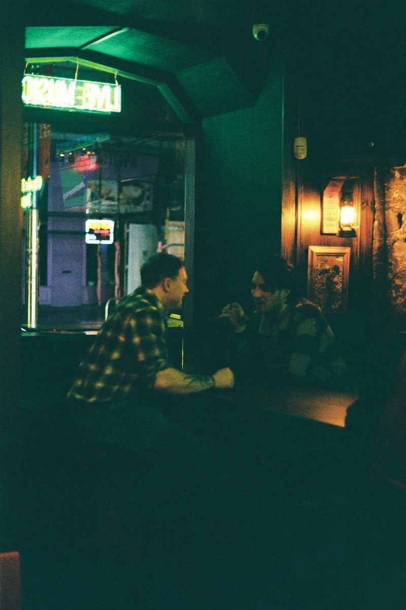 Two people sitting across from each other in a dimly lit bar, engaged in conversation, with neon signs and framed pictures on dark-colored walls in the background.