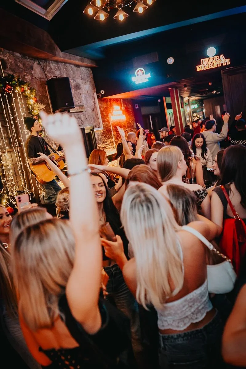 Crowd of people dancing and enjoying live music in a bar with Christmas decorations, a singer playing guitar, and neon signs on the walls.